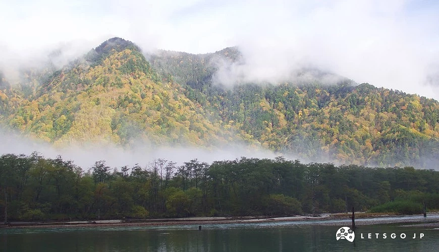 煙霧圍繞在金黃色的山脈上,前面有綠色樹林跟一條溪流