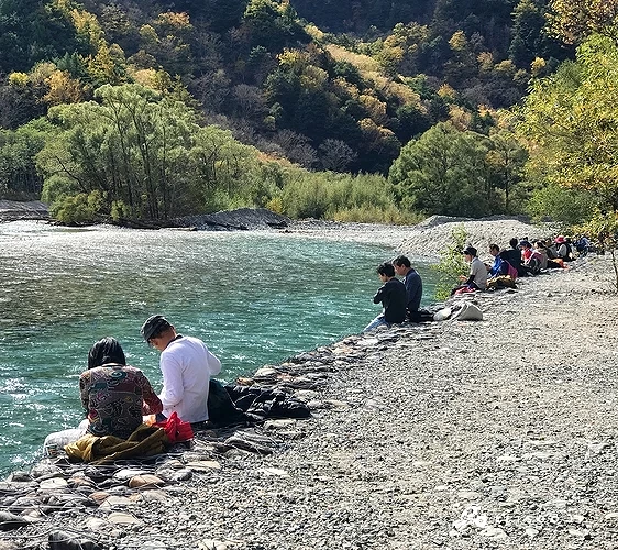 後面是綠色樹林和山脈,左側有藍綠色溪流,右側石子岸邊有一排人坐著
