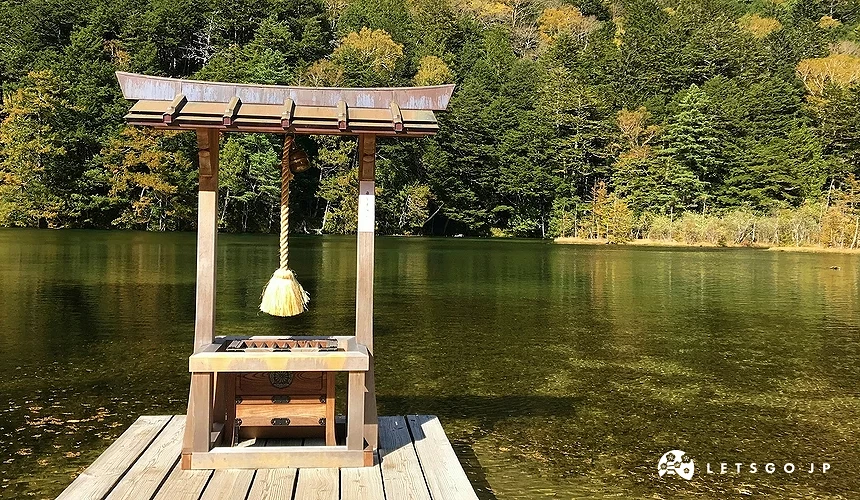 後面是黃綠色樹林,前面有一片湖,湖的前面是一座木橋和木製小神社