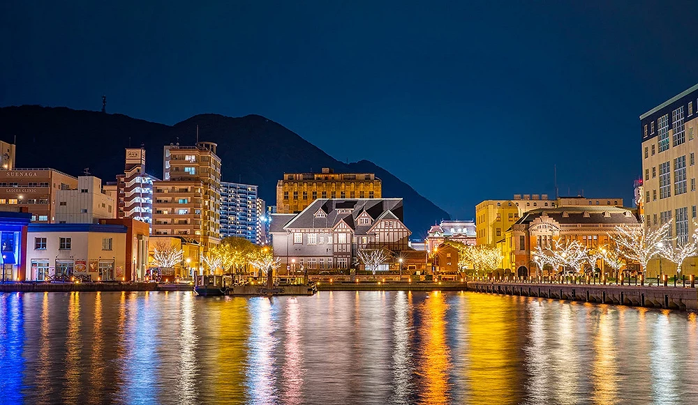 北九州門司港夜景全景,燈飾街道與水面倒影交織,洋風建築群在山景背景中閃耀