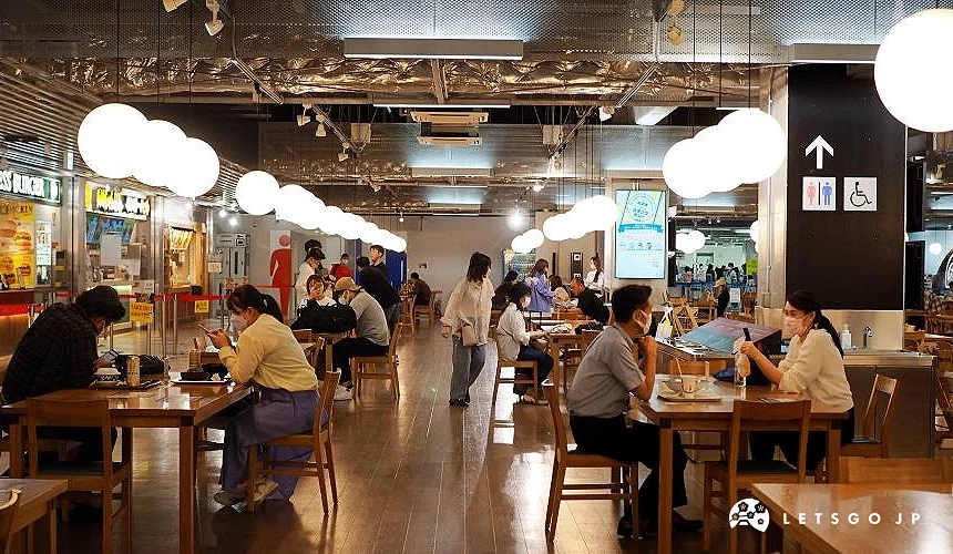成田機場第三航廈2樓的美食廣場,旅客坐在木質桌椅區用餐,後方可見各式餐廳與明亮的燈飾,氣氛熱鬧舒適