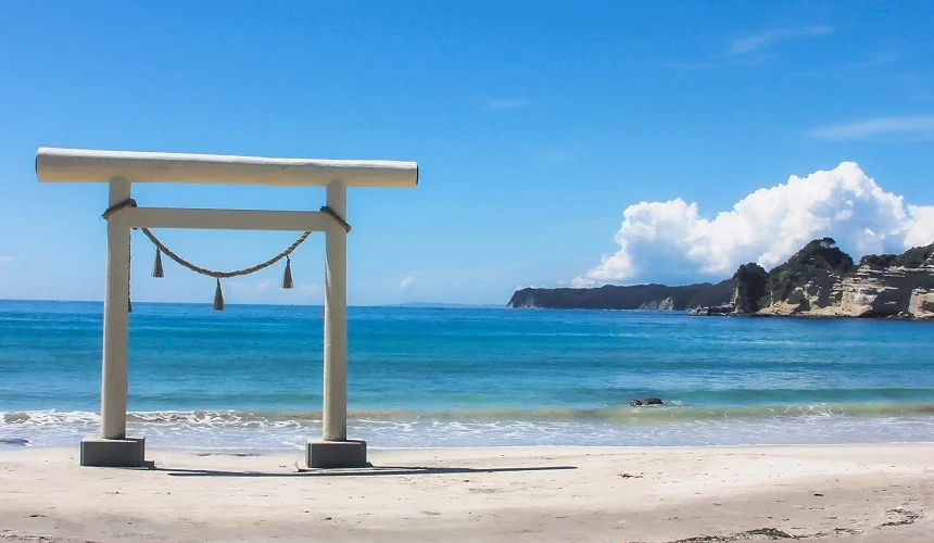 白色鳥居矗立在沙灘上,背後是湛藍大海與遠方海岸。