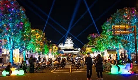 姬路城前聖誕節夜景,街道兩側樹木點亮多彩燈飾,天空中有藍色光束照向城堡,街中央設有攤位與座椅,人潮熱鬧