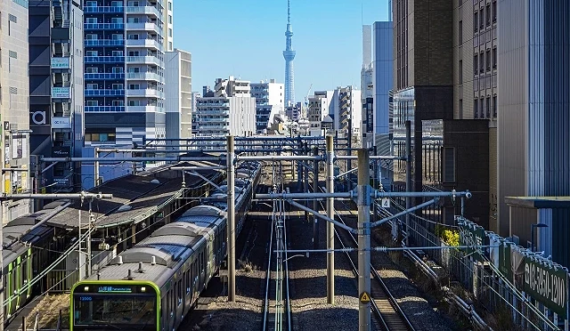 行駛中的 JR 山手線列車穿梭於東京市區鐵道上,可見遠方的晴空塔,呈現東京典型城市交通景象