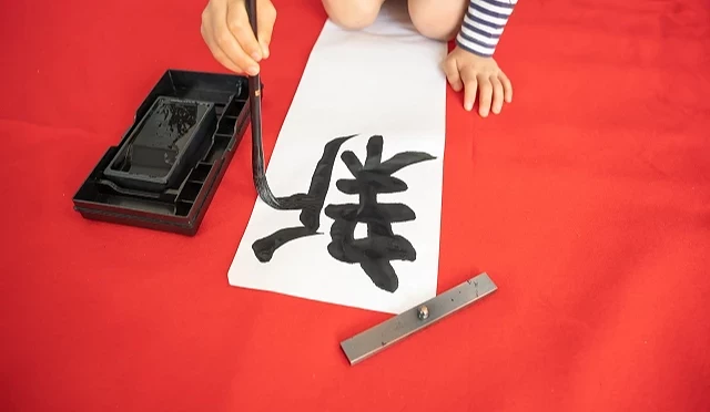 孩童在紅色墊布上以毛筆書寫新年開筆作品,白紙上寫有大大的黑色漢字,呈現日本新年開筆的傳統場景