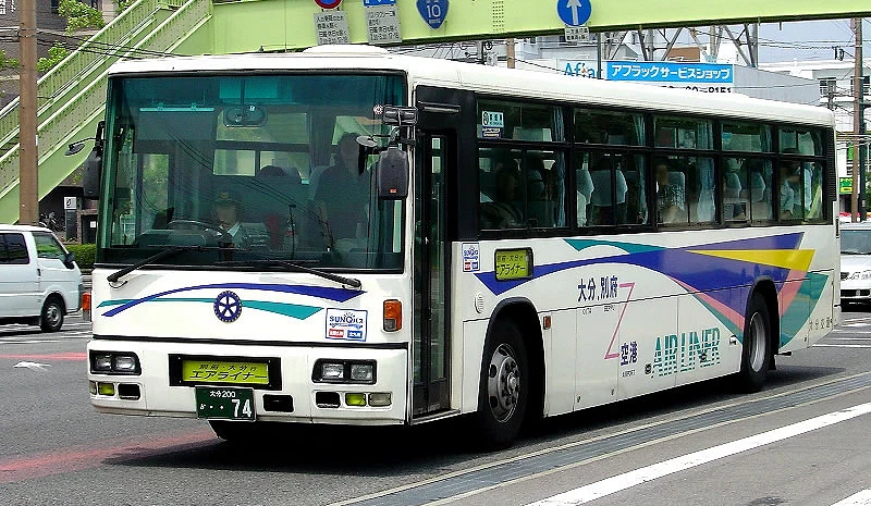 一輛往返大分機場與市區的空港特急巴士「Airliner」行駛在市區道路上,白底車身配有藍綠色與紫色線條,前方可見斑馬線與綠色天橋