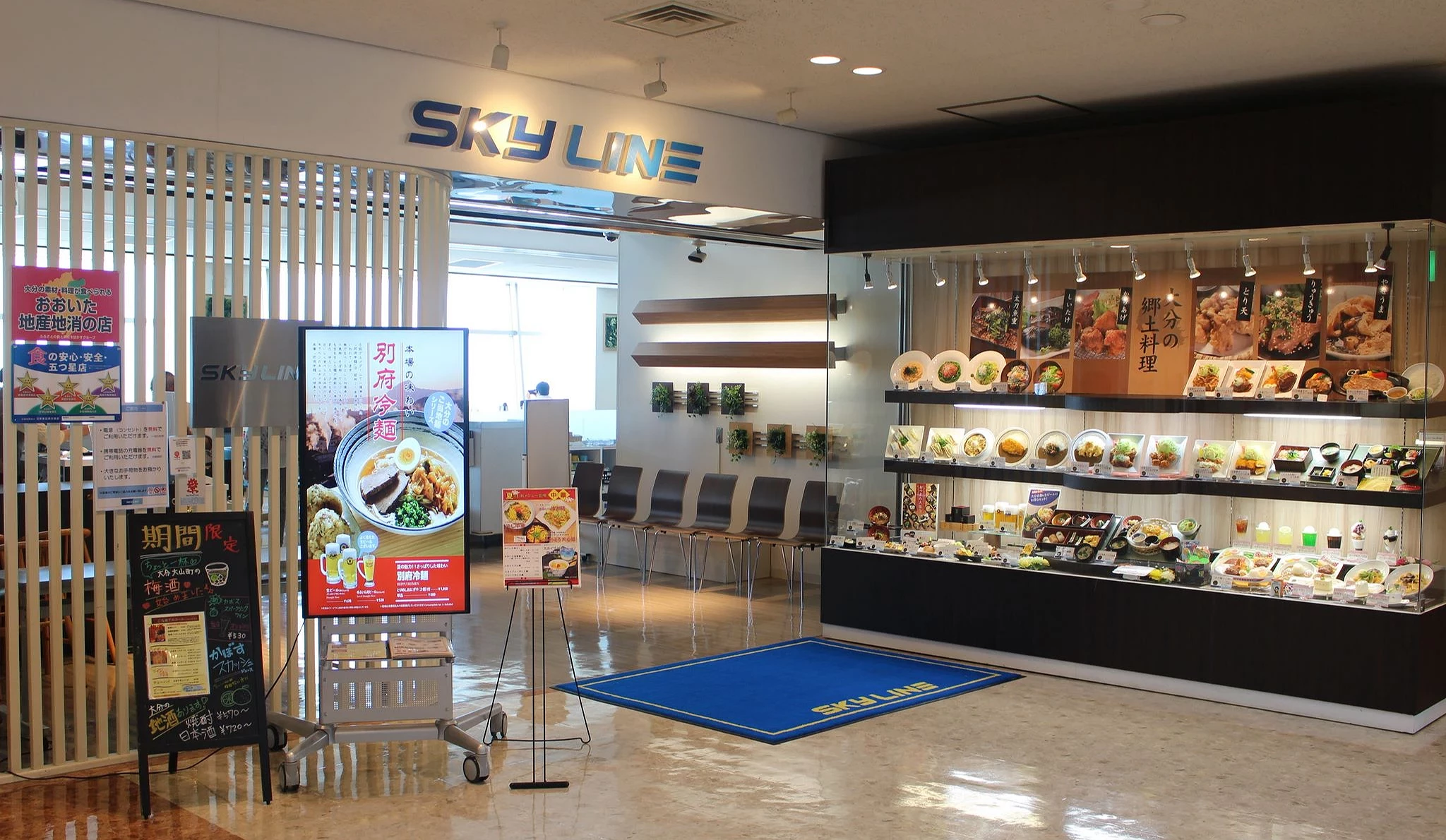 大分機場三樓「View Restaurant SKY LINE」店鋪入口,可見料理模型櫃展示多款大分名物,如炸雞天婦羅、豐後牛與各式定食,入口區旁設有告示牌與電子看板,提供餐點介紹與期間限定資訊