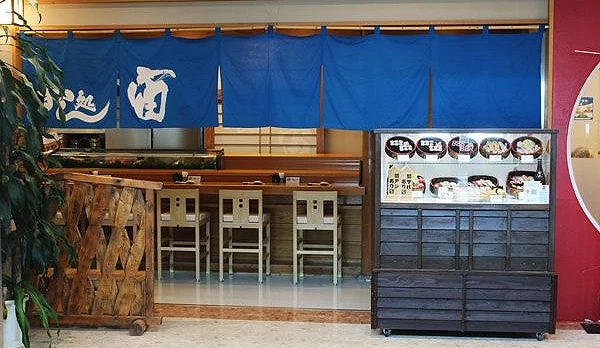 大分機場三樓壽司店「Sushi Restaurant SORA」入口,藍色暖簾下方是木製吧台座席,右側展示櫃內陳列多種握壽司與海鮮丼餐點模型