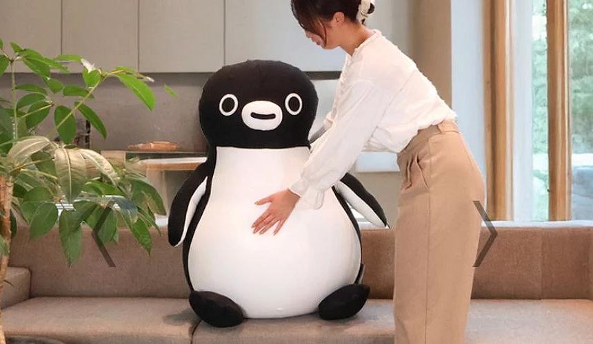 一位女性正在沙發旁抱著等身大的 Suica 企鵝抱枕,玩偶體型巨大、外觀圓滾可愛