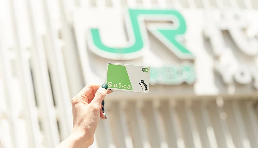 一隻手在 JR 車站建築前舉起 Suica 卡,背景為模糊的 JR 標誌,卡面清晰呈現綠色設計與企鵝插圖