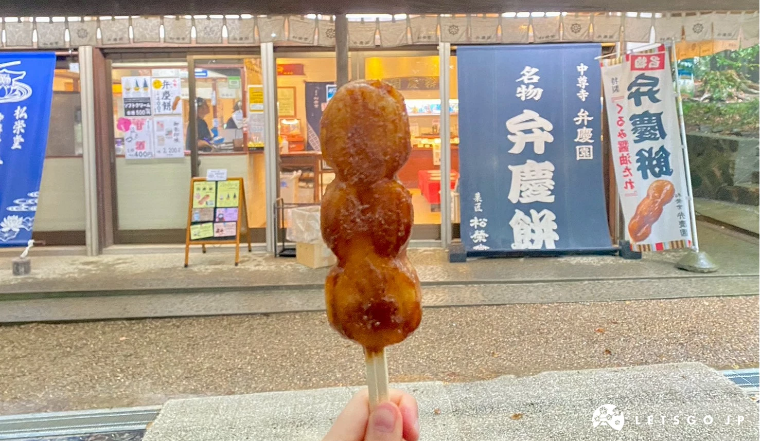 一隻手拿著弁慶餅,背景是松榮堂弁慶園