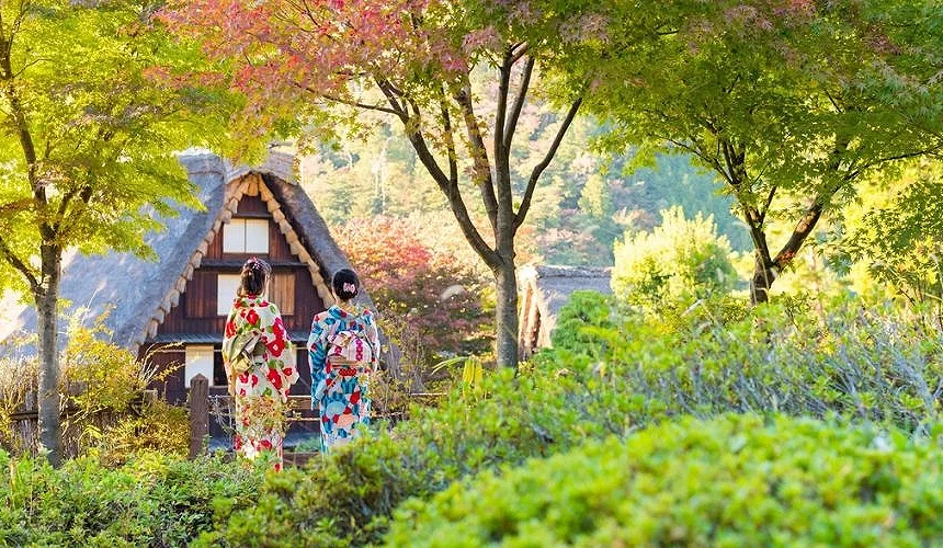 兩位穿著和服的旅客走在下呂溫泉合掌村的園區中,周圍綠意與紅葉交織,背景可見茅草屋