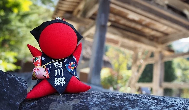 紅色的白川鄉吉祥物「さるぼぼ」布偶放在石頭上,背景為白川鄉傳統合掌屋建築
