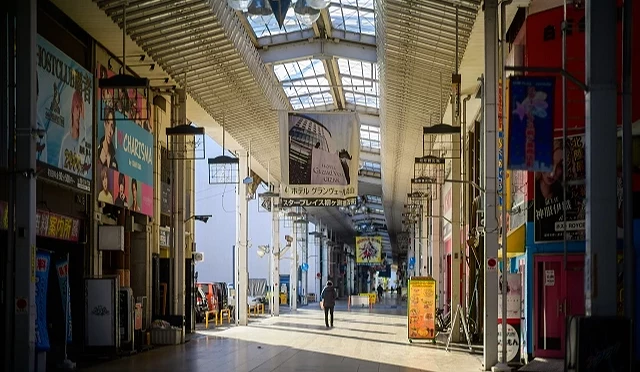 岐阜市區拱廊商店街的白天景象,陽光灑落在地面,一名行人走在中央,兩側可見各式店家與色彩鮮明的招牌