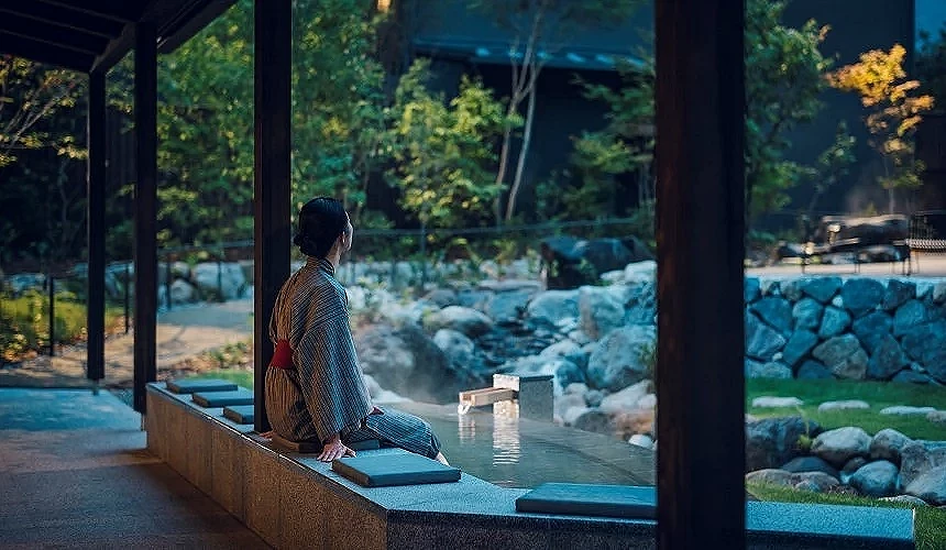 浴衣旅客坐在半露天足湯邊緣,面向佈置有岩石與綠意的日式庭園,蒸氣輕飄
