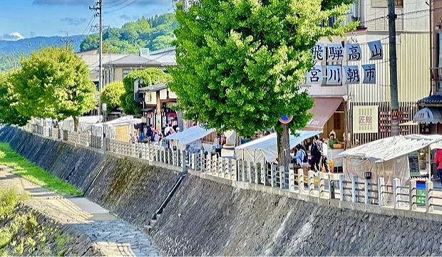 高山宮川朝市的河畔市集景色,攤販帳篷排列在步道旁,旅客在蔭影與藍天下悠閒逛市集