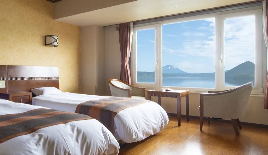 洞爺湖溫泉飯店湖景雙床客房,窗外可見洞爺湖與羊蹄山,房內配置桌椅與大片窗景,適合欣賞湖景與花火