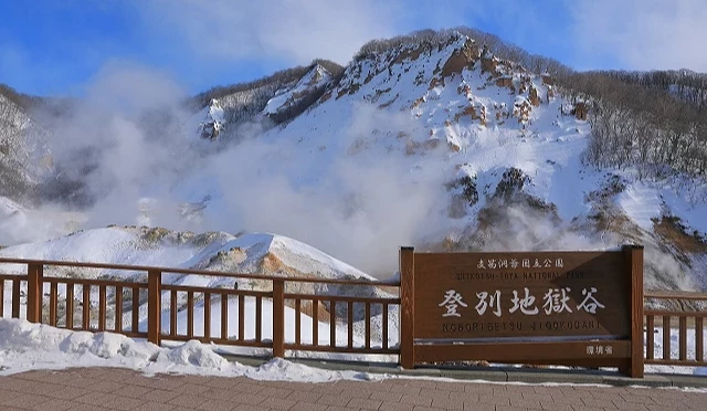 冬季登別地獄谷景色,雪覆火山岩壁間冒出白色蒸氣,前方設有登別地獄谷木製標誌與觀景步道
