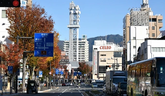 甲府市市區街景,主要道路兩側林立商店與百貨大樓,背景可見市區地標建築與山景