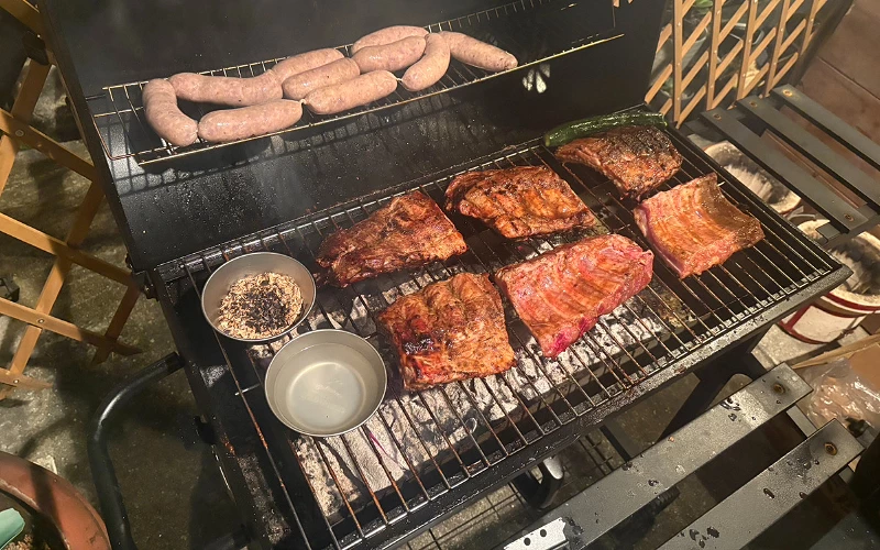 烤爐上燒烤牛肋排與香腸,旁有調味碗與蔬菜