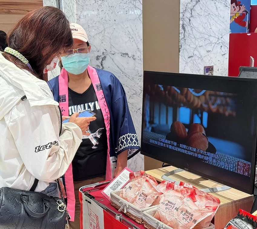 工作人員與顧客交流,櫃台上放有產品型錄與樣品,後方螢幕展示著美味的水蜜桃特寫。
