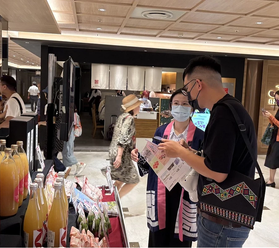物產展現場工作人員正在向顧客介紹產品,背景可見各式飲品展示。