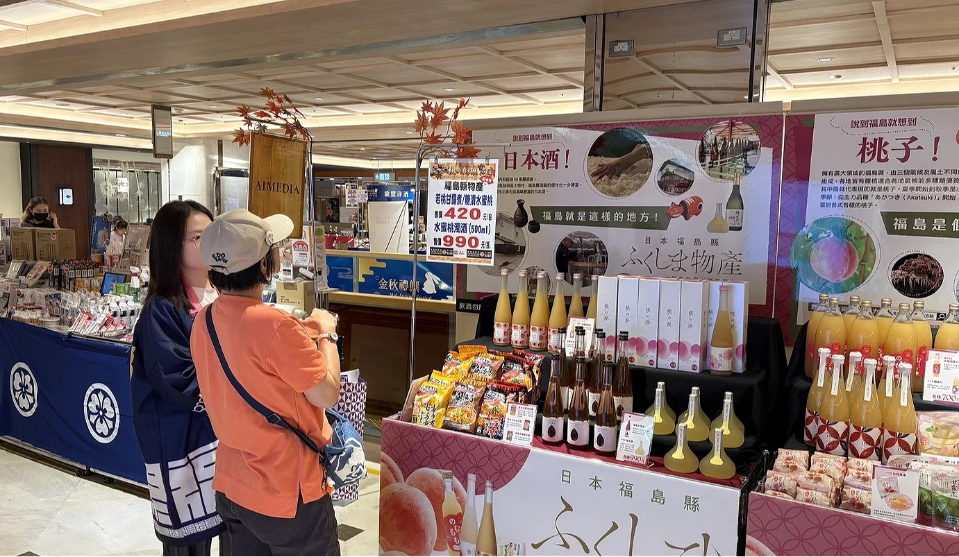 福島物產展位全景,身穿法被的工作人員正與顧客交談,後方背板印有巨大的水蜜桃與清酒文宣。