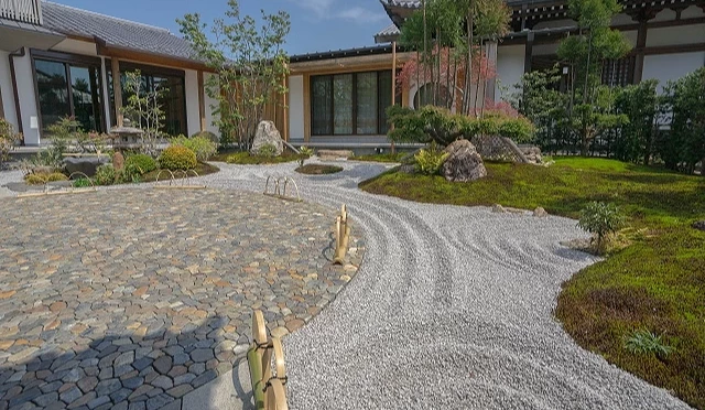 精緻的白砂紋路與圓潤石塊鋪設的地面,其間點綴著翠綠苔蘚坡地、修剪整齊的植栽與一座石燈籠。背景為日式傳統建築與木製迴廊