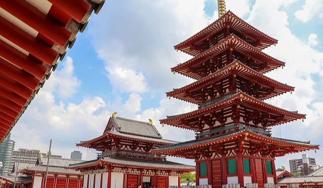 大阪四天王寺五重塔的仰角景觀,朱紅色多層屋簷與金色塔剎在藍天白雲下映照,左側背景可見金堂屋頂