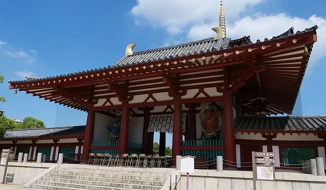 大阪四天王寺中心伽藍的「中門」(仁王門)正面外觀,朱紅色傳統建築搭配深色瓦頂與金色鴟尾,門洞兩側可見巨大的仁王像,前方有石階與碎石地