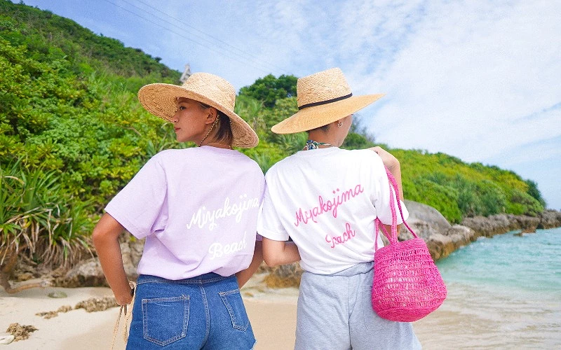 兩名女子背對鏡頭站在海邊,上衣背面印有 Miyakojima Beach 字樣