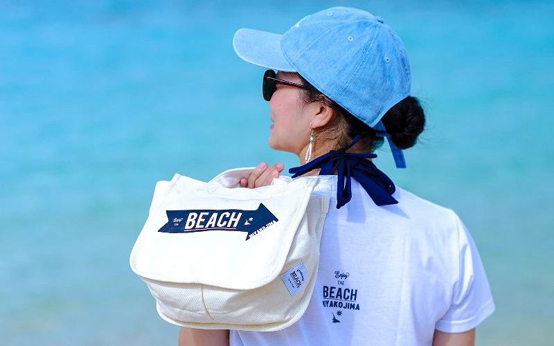 女子背對鏡頭站在海邊,肩背印有 BEACH 字樣白色帆布包