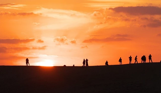 鳥取砂丘夕陽景色,橘紅色天空下多名遊客剪影站在沙丘稜線上,夕陽映照出壯闊日落畫面。