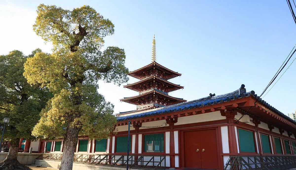 大阪四天王寺的五重塔,朱紅色塔身聳立於迴廊後方,左側有茂密的大樹襯托,背景為晴朗藍天