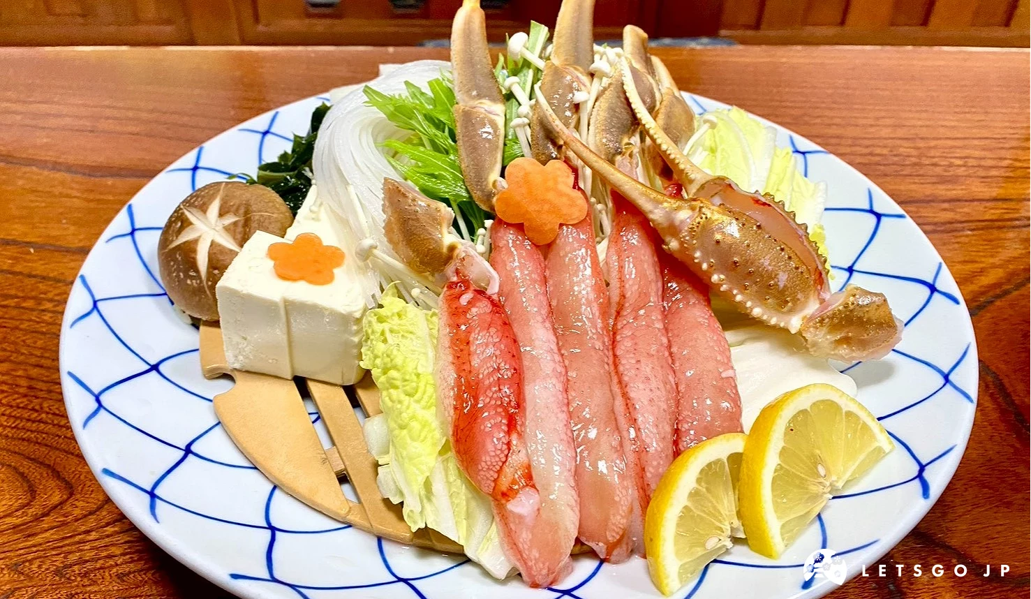 一張特寫照片,白藍花紋的圓盤上整齊擺放著多支肉質紅潤、已去殼的長蟹腳,旁邊配有新鮮白菜、豆腐、香菇、金針菇與檸檬片。