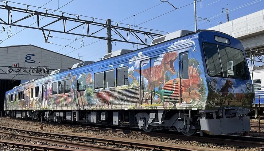 藍色電車車身繪有彩色恐龍壁畫,停靠於鐵軌車廠,寫有日文標語。