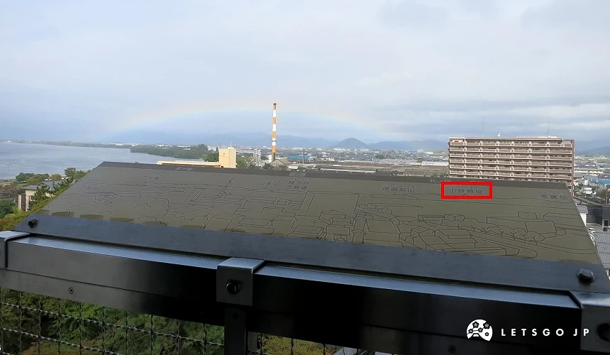 城閣欄杆上刻有城鎮地圖的導覽板,紅框標示「小谷城址」,遠方可見彩虹與湖景