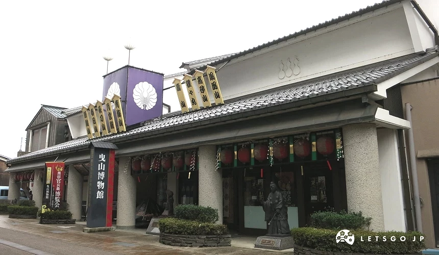 曳山博物館外觀,上方掛有豐臣五七桐紋紫色大旗,入口前設有人物銅像