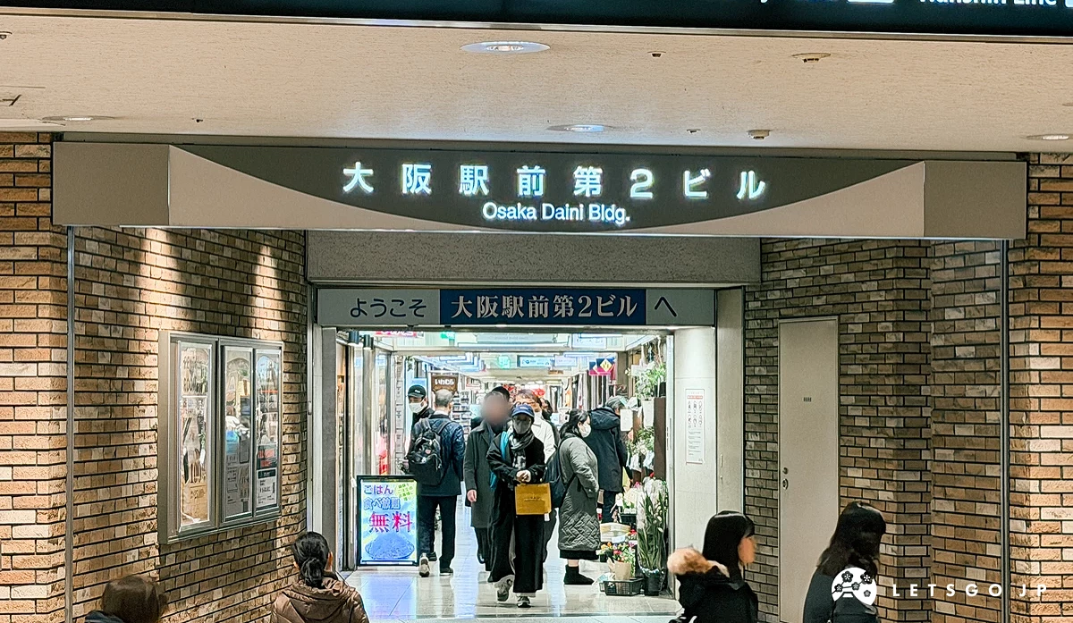 大阪站前第 2 大樓的地下入口。上方有咖啡色橫向招牌,寫著白色漢字「大阪駅前第 2 ビル」。下方為玻璃自動門,門內有穿著深色衣服的行人經過