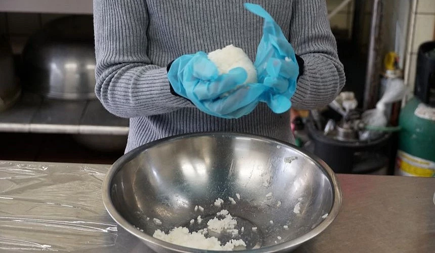 一名穿著灰色毛衣、戴著藍色塑膠手套的人,正在鋼盆上方揉捏一顆渾圓潔白的飯糰。