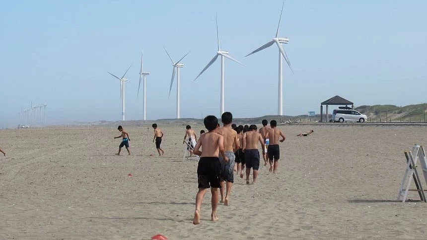 廣闊的沙灘上,一群人正朝著遠方巨大的白色風力發電機跑去,背景是清澈的藍天。