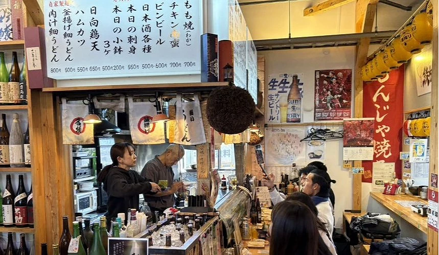 熱鬧的居酒屋內部。暖色調燈光下,吧檯內廚師正在忙碌,架上擺滿日式酒瓶,充滿生活感。