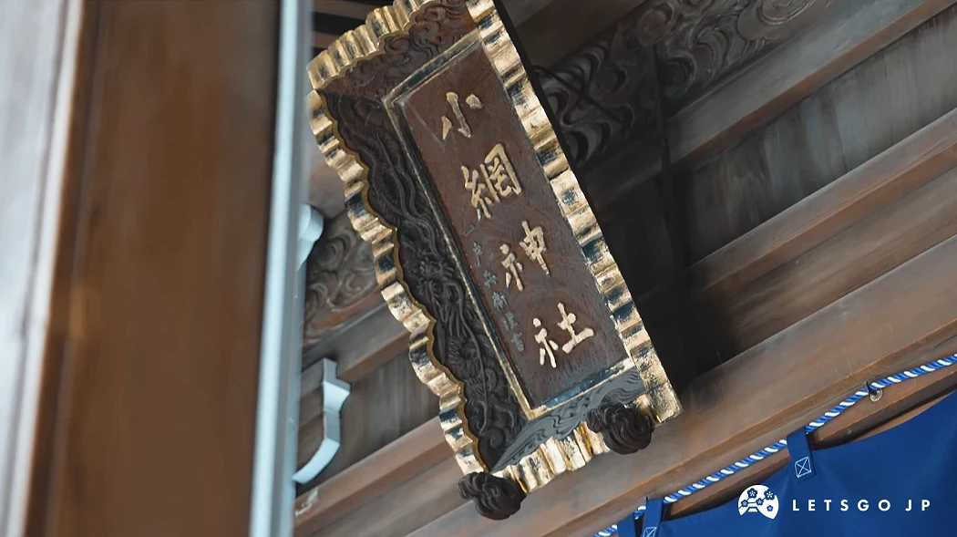 小網神社的木造牌匾特寫。金色邊框的黑色牌匾上刻著「小網神社」金色字體,背景為層次分明的木造屋頂結構與深藍色布幔。