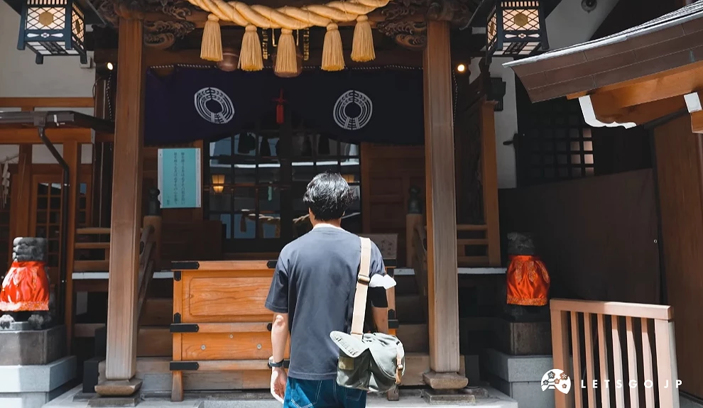 一位背著背包的男士正面向木造的本殿建築。畫面中央可見木製賽錢箱,兩側有石燈籠與掛著紅色布幔的裝飾,展現日本下町神社的日常參拜景象。