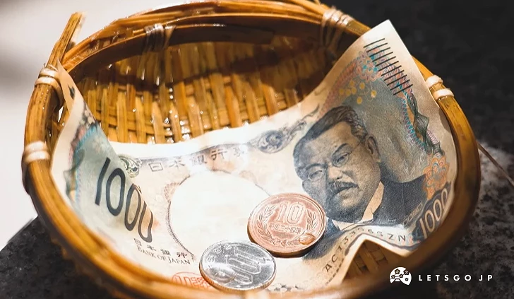 竹編小簍子的近景特寫,裡面裝有一張一千日圓紙鈔及數枚硬幣,放置在洗錢池旁的石頭上。