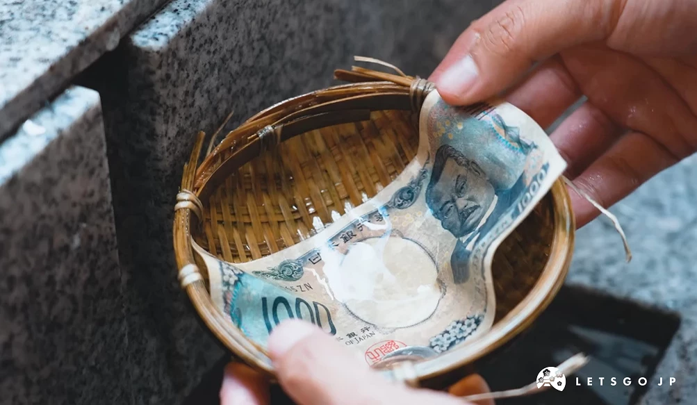 畫面捕捉到正在洗錢的動態。一隻手扶著裝有錢幣與紙鈔的竹簍浸入水中,另一隻手正拿著紙鈔,池水清澈見底。