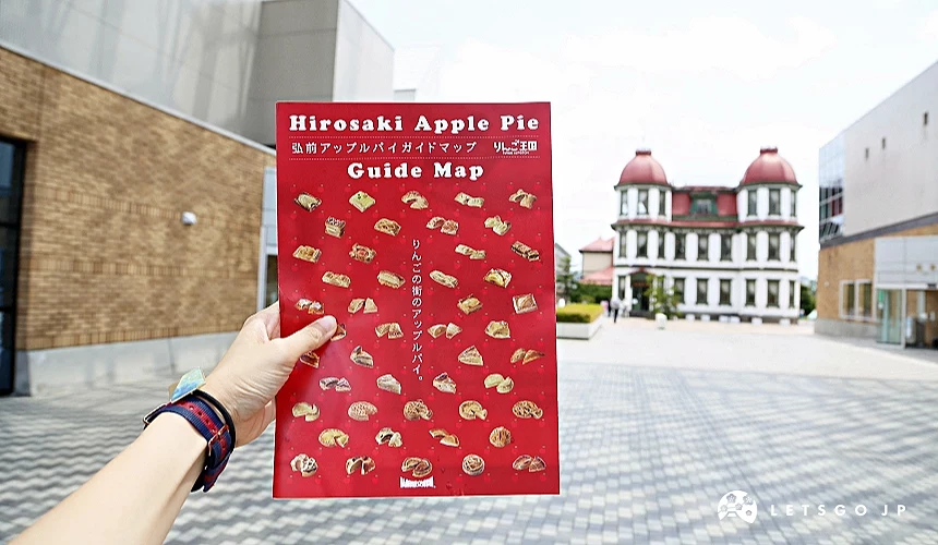 手持紅色弘前蘋果派指南地圖,背景為弘前市街景與歷史建築,地圖上標示多款蘋果派圖示