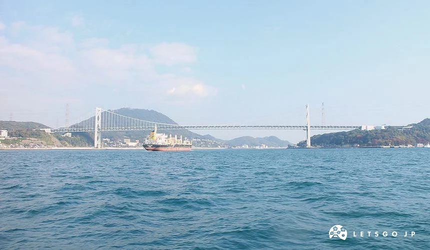 關門海峽海面與關門橋景觀,一艘貨船航行於橋下,遠方為山景