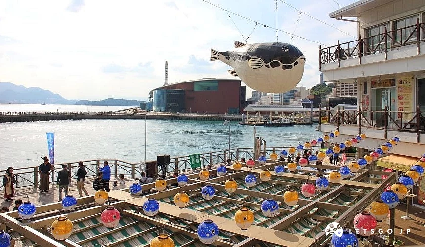 唐戶市場附近海港景色,空中懸掛河豚裝置,水岸步道與遊客活動場景