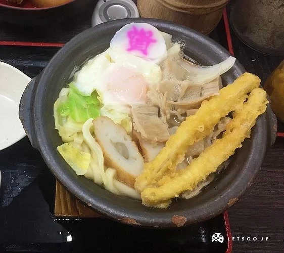 一碗日式麵食料理,上方有溫泉蛋與天婦羅,盛裝於黑色鍋中